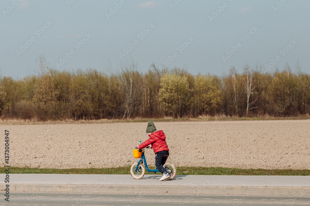 Obraz premium A child in a red jacket and green hat rides a blue balance bike on a paved path next to a field. The child is facing away from the camera, and the field in the background is brown and barren.