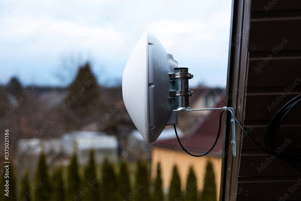 A white WiFi router dish is mounted on a house wall. The background shows rooftops, trees, and tall shrubs under an overcast sky in a suburban area.