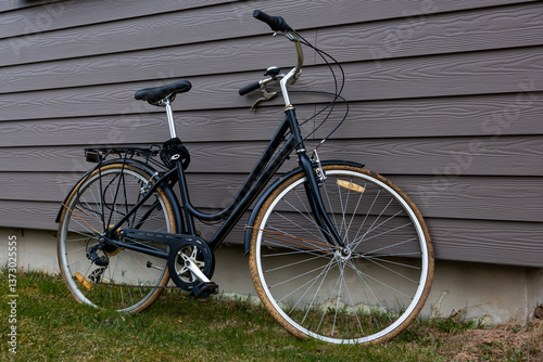 A black bicycle with a step through frame leans against a gray wooden house wall. Grass and soil are visible at the base, suggesting a residential setting.