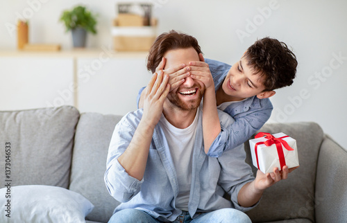 Happy father's day. Son congratulating his daddy, covering his eyes from behind, greeting and giving present at home. Family holiday celebration, surprise present for dad