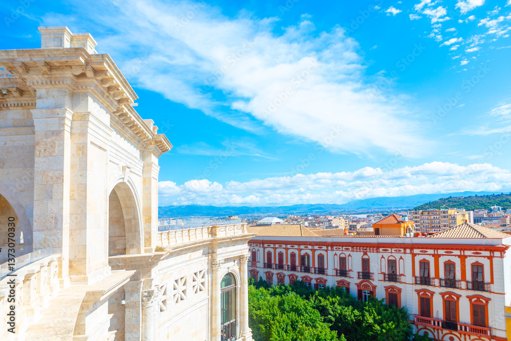 Sunlit view of Bastione di Saint Remy in Cagliari, Sardinia, limestone arches against a vivid blue sky. Historic architecture contrasts with lush greenery and an elegant neoclassical building below