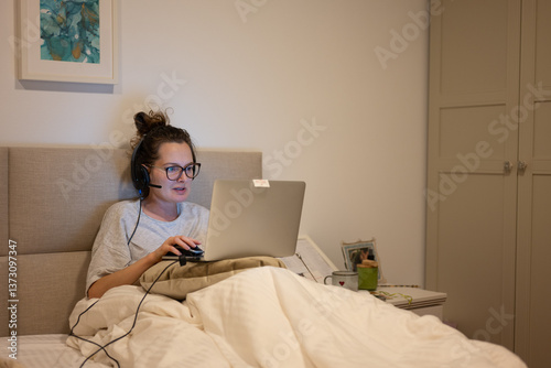 Young woman wearing headset working from home using laptop in bed, early in the morning or late at night