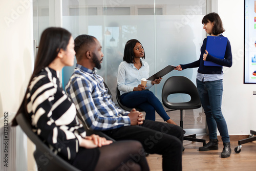 HR female representative invites black candidate to job interview, holding her resume in company waiting area. African american woman giving her application to caucasian recruiter in office lobby.