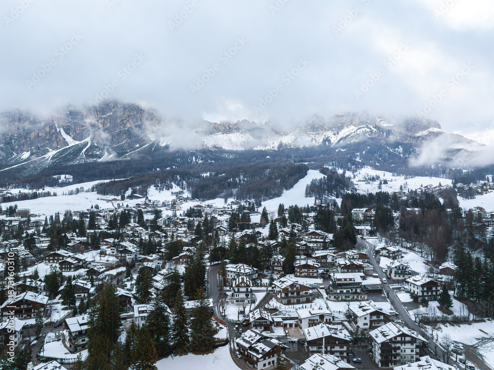 Aerial view of Cortina d'Ampezzo in Italy, featuring snow covered alpine architecture, dense forests, ski runs, and Dolomites peaks under cloudy skies.