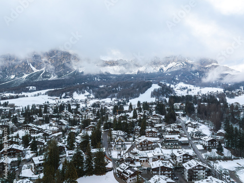 Obraz na płótnie Aerial view of Cortina d'Ampezzo in Italy, featuring snow covered alpine architecture, dense forests, ski runs, and Dolomites peaks under cloudy skies. w salonie