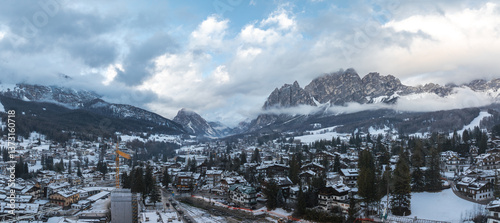 Fotografie Aerial view of Cortina d'Ampezzo, Italy, showing snow covered rooftops, alpine buildings, forested valleys, mountain peaks, and construction activity