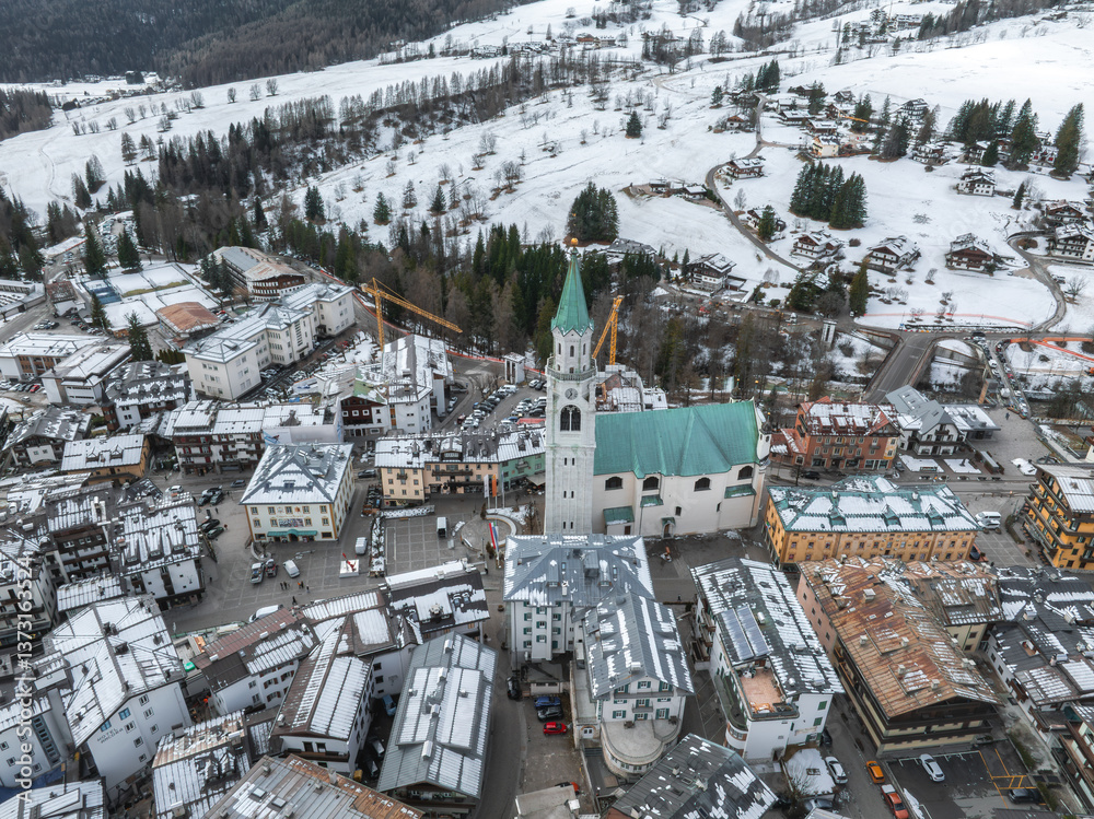 Aerial view of Cortina d'Ampezzo, Italy, featuring a green roofed church, snow dusted buildings, alpine slopes, and construction cranes in the background.