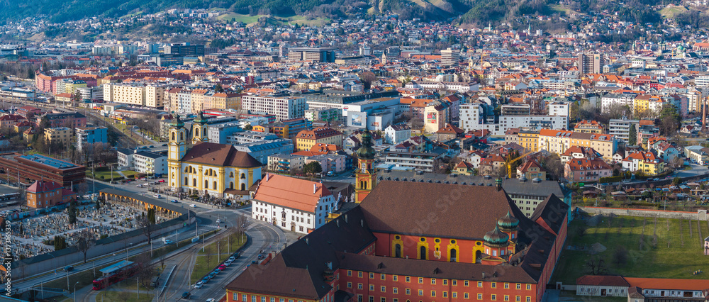 Aerial view of Innsbruck, Austria, featuring Wilten Basilica with twin yellow towers, a cemetery, historic buildings, and the Nordkette mountain range.