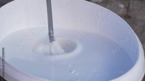 Plaster mixer or universal tool lowers into container with white paint, stirring for even texture. This step prepares paint for smooth application on walls during home or building construction