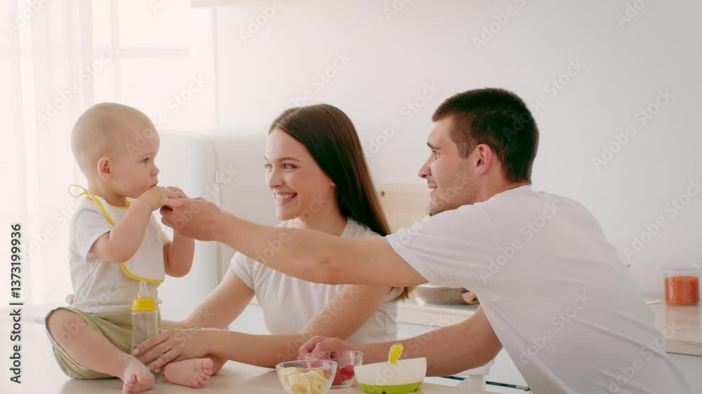 In a bright kitchen, a joyful toddler is laughing while holding a snack. Both parents are engaged, smiling and sharing a loving moment during mealtime at home.