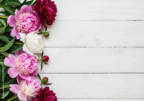 Elegant pink, red, and white peonies with lush green leaves arranged on a white wood background.