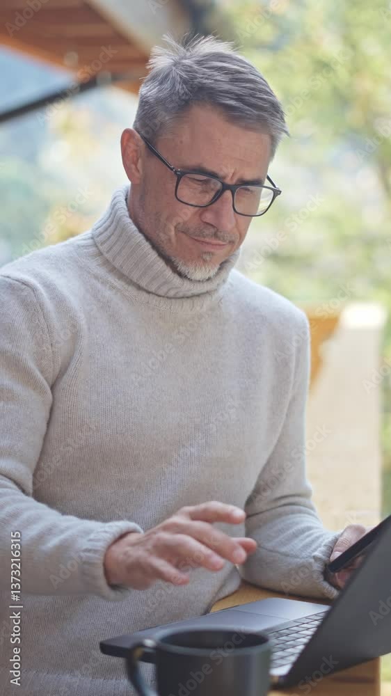 Smiling mature businessman holding smartphone and using laptop while working remotely on wooden terrace. Entrepreneur managing business from home office. Portrait of smiling satisfied mid adult man. Smiling mature businessman holding smartphone and using laptop while working remotely on wooden terrace. Entrepreneur managing business from home office. Portrait of smiling satisfied mid adult man.