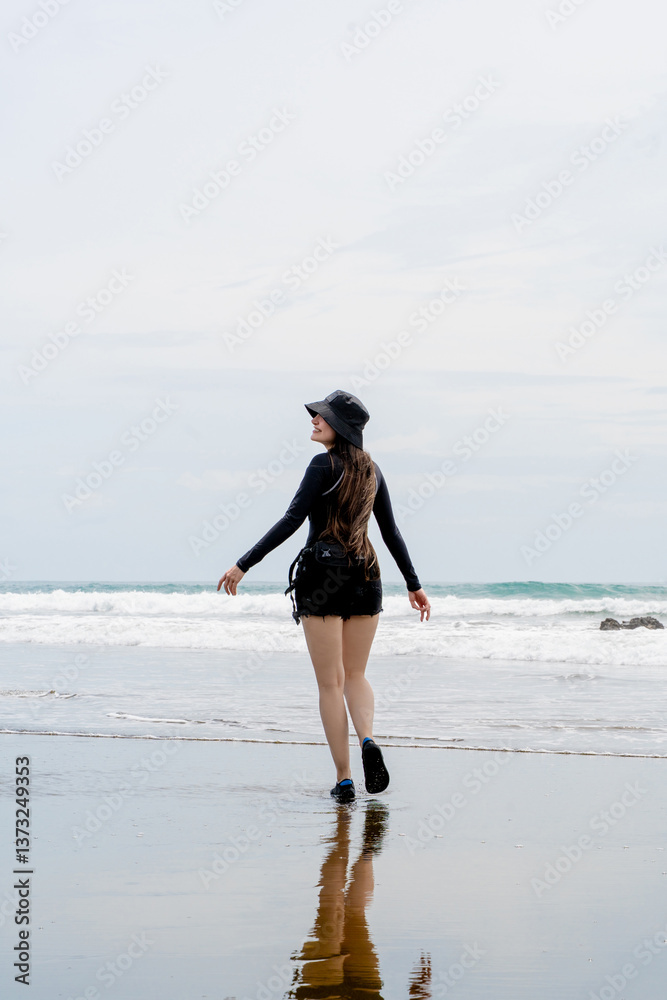 Happy young woman with long hair and a black hat walks along a beach, with her reflection visible on the wet sand. The peaceful coastal setting enhances the moment. Happy young woman with long hair and a black hat walks along a beach, with her reflection visible on the wet sand. The peaceful coastal setting enhances the moment.