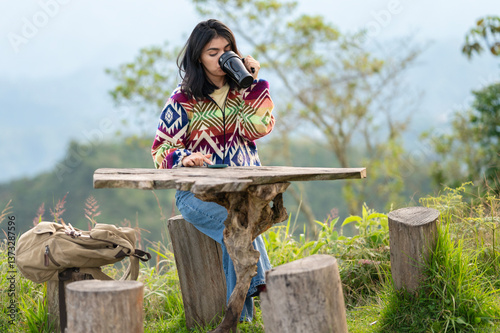 Woman drinking from reusable mug and browsing smartphone in mountain scenery
