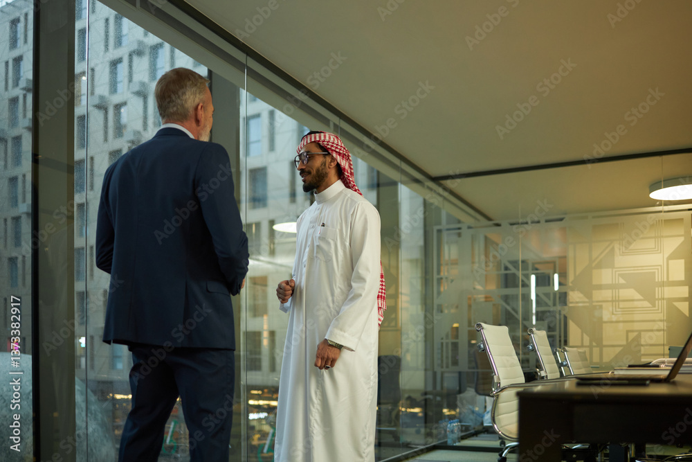 Medium long shot of mature man in formal suit and his Middle Eastern business partner standing in modern office and talking Medium long shot of mature man in formal suit and his Middle Eastern business partner standing in modern office and talking