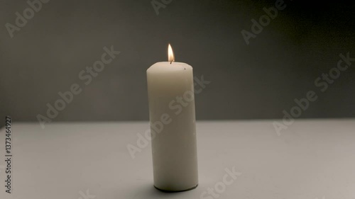 A close-up of a long white burning candle standing in the middle of a table in a dimly lit room. Peace and tranquility from a burning light