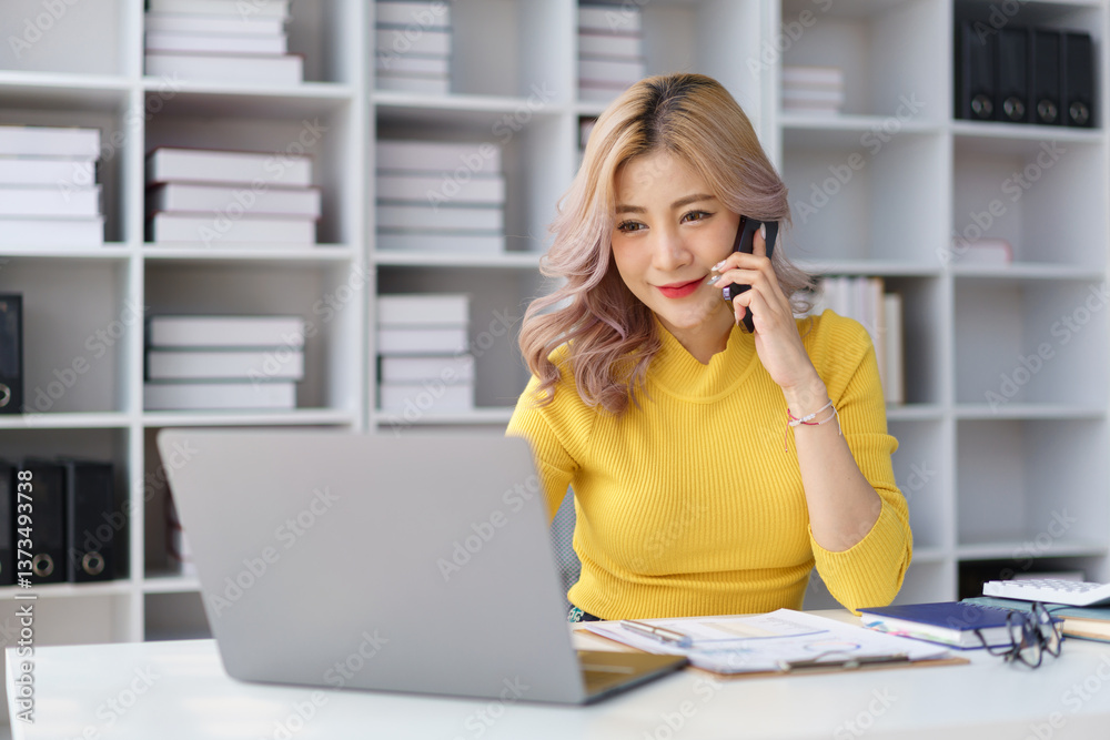 Confident businesswoman talking on mobile phone while working on laptop at modern office desk.. Confident businesswoman talking on mobile phone while working on laptop at modern office desk..