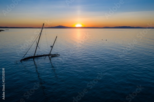 A sunken sailboat rests in calm waters at sunset in San Pablo Bay, Rodeo, California, USA. The boat's mast extends above the surface, a reminder of past voyages. The sun reflects on the water.