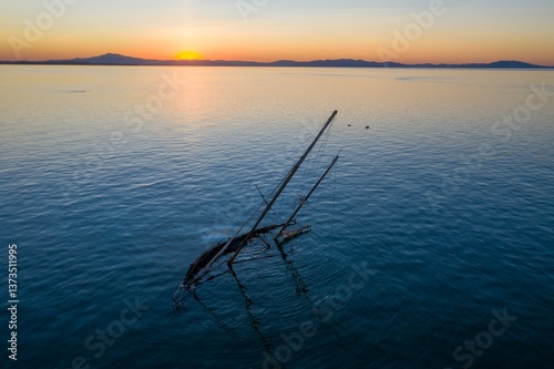 A sunken sailboat sits in San Pablo Bay, Rodeo, California, USA, as the sun rises over the distant mountains. The boat's masts point skyward, a reminder of past voyages.