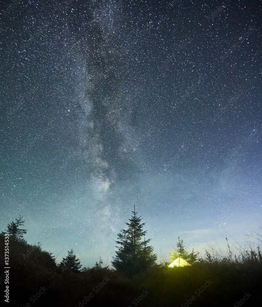Fototapeta premium Radiant yellow tent beneath awe-inspiring, star-filled sky with Milky Way prominently stretching across. Surrounded by pine trees and silhouetted by hill, scene epitomizes tranquility of nature.