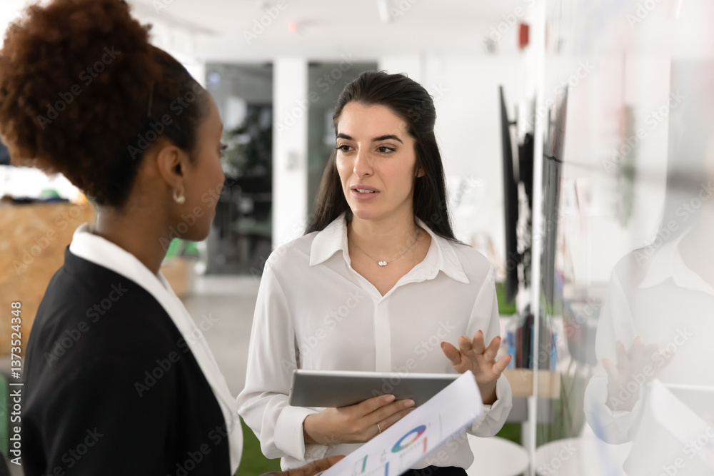 Fototapeta premium Serious young Latin project manager woman talking to female African colleague in office training education space, standing at whiteboard, speaking, holding digital tablet