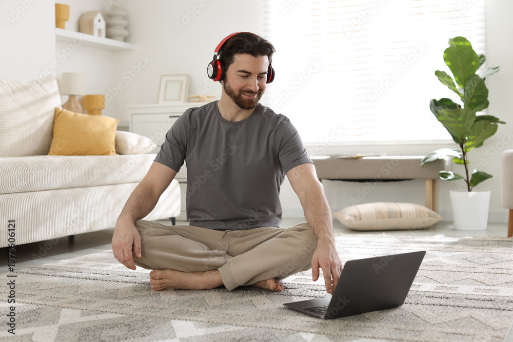 Smiling man in headphones meditating near laptop on floor at home