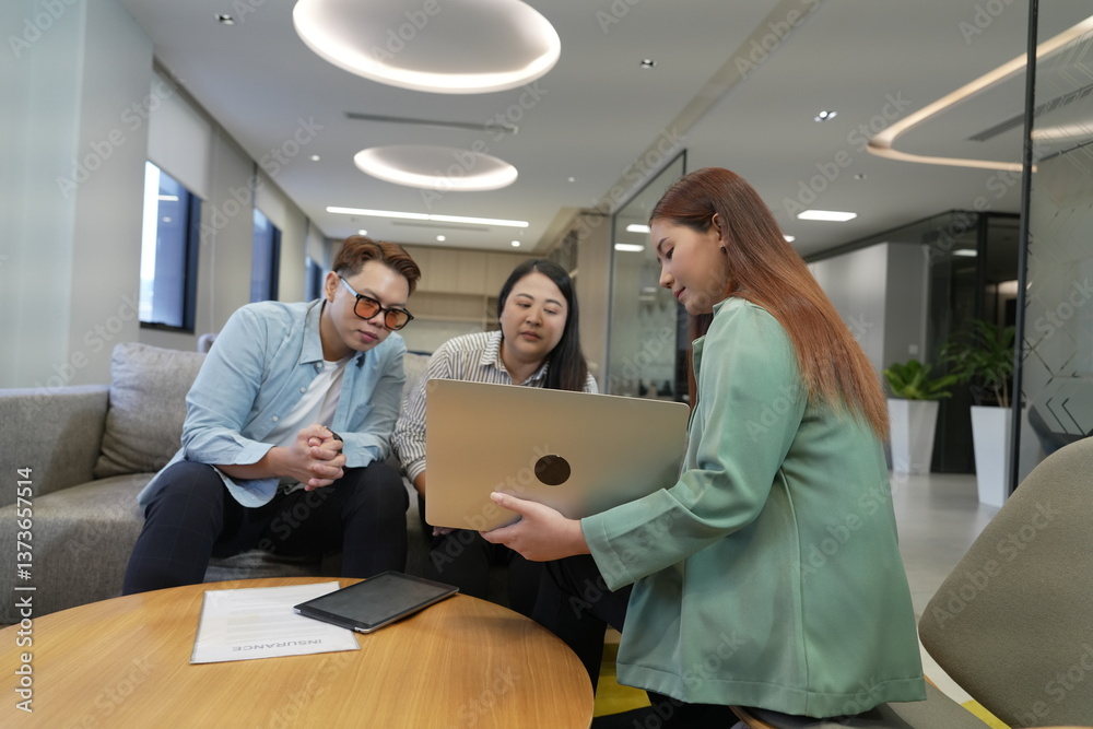 A 29-year-old Asian woman in a suit holds a laptop explaining the terms and conditions of life insurance to male and female customers in an office lounge.