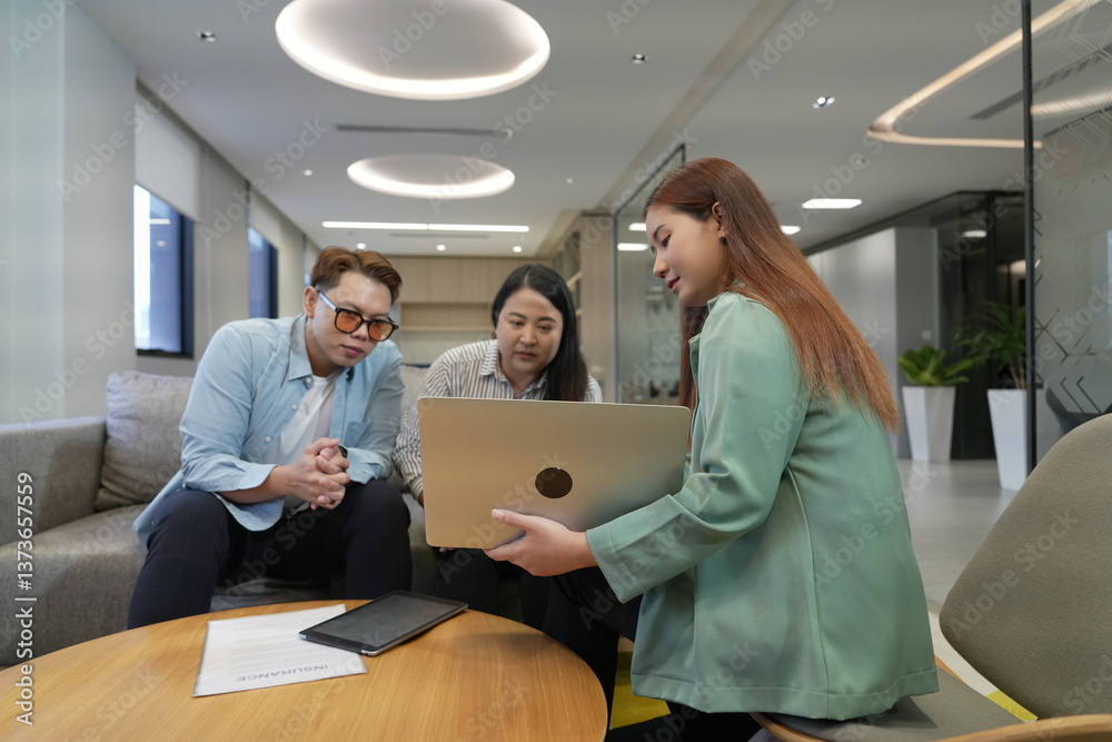 A 29-year-old Asian woman in a suit holds a laptop explaining the terms and conditions of life insurance to male and female customers in an office lounge.