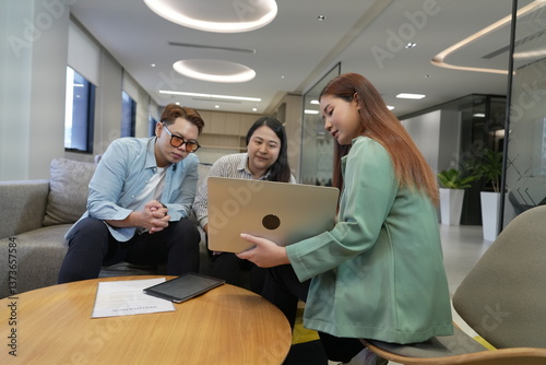 A 29-year-old Asian woman in a suit holds a laptop explaining the terms and conditions of life insurance to male and female customers in an office lounge.