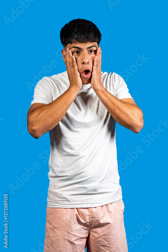 Quadro su tela Shocked young man holding face in disbelief on blue background