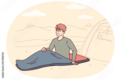 Man hiker with sleeping bag sits on ground on camping site near tent located in national natural park. Smiling guy traveler waking up in morning and spending time in camp for camping
