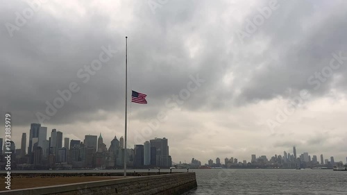 American flag at half-mast waves in foreground with New York City skyline in distance under dramatic cloudy sky, for patriotic ads, travel promos, and urban visuals, USA
