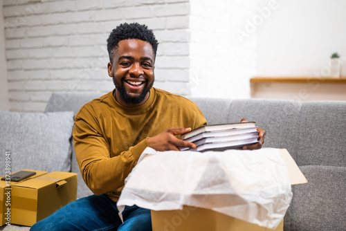 Happy young African American man smiling and opening delivered package with books while sitting on sofa at his home. 