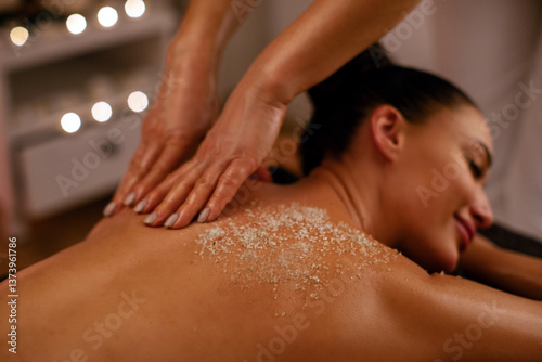 Lady deeply relaxing while spa professional applying sea salt, exfoliating treatment to woman's back, promoting wellness and skin health