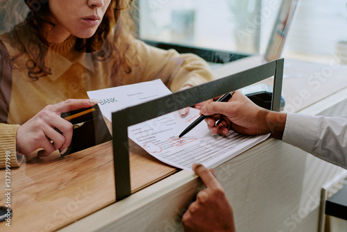 Detailed view of person filling out forms at bank counter, interacting with bank teller behind glass divider in background
