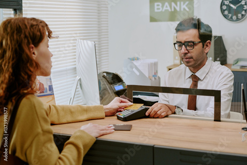 Customer engaging with bank teller during transaction in modern bank setting, handing over card for service, office environment visible in background including computer and documents