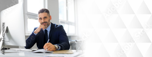 Portrait of happy middle aged man entrepreneur working on computer and taking notes, sitting at workplace and smiling at camera, writing something to notepad, sitting at workplace, copy space