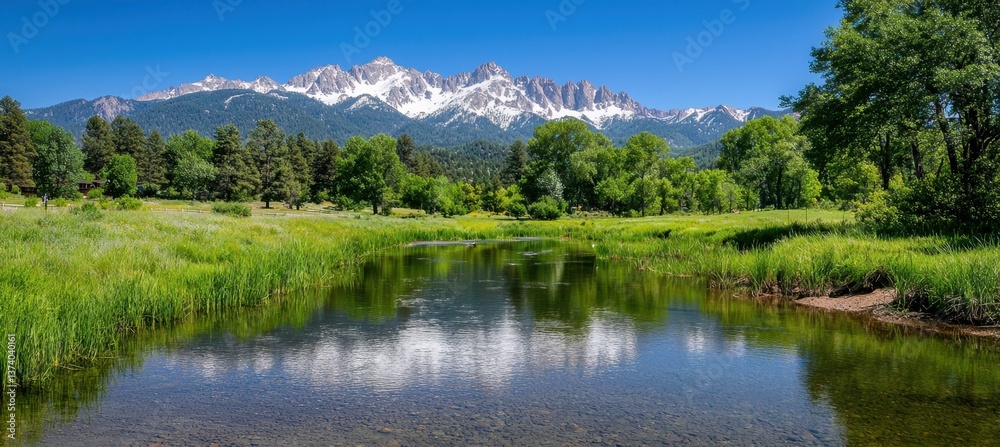 Fototapeta premium A serene landscape featuring a river bordered by lush greenery, with majestic snow-capped mountains under a clear blue sky in the background.