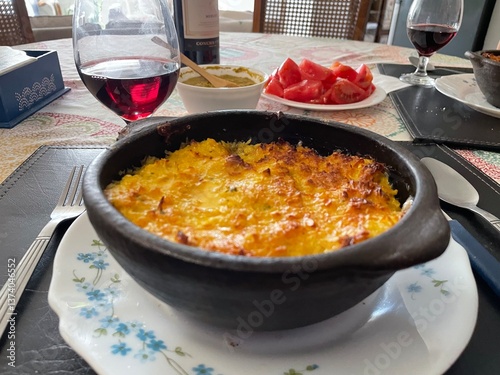 Pastel de choclo, platillo típico chileno en un librillo de greda, etá en una mesa bien servida junto a vino tinto y tomates. photo