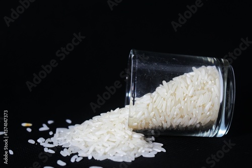 A clear glass tilted sideways against a solid black background. The glass is cylindrical, has a smooth, reflective surface, and contains some raw white rice grains. The rice flows out of the glass.