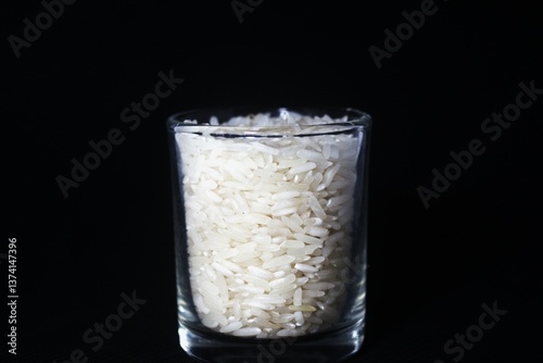 A clear shot glass filled with raw white rice grains. The shot glass is placed in the center against a solid black background, creating a strong contrast that emphasizes the texture and color of the r