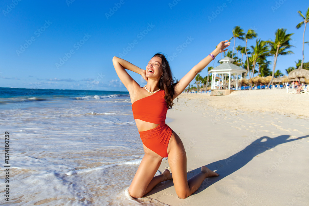 Fototapeta premium Happy woman in swimsuit enjoying sun and wind, posing at the edge of ocean surf on the beach of tropical resort