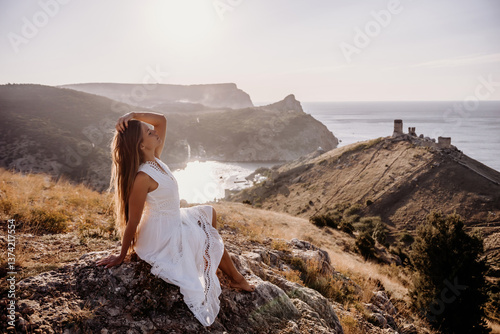 A woman in a white dress is sitting on a rock overlooking a body of water. She is enjoying the view and taking in the scenery.