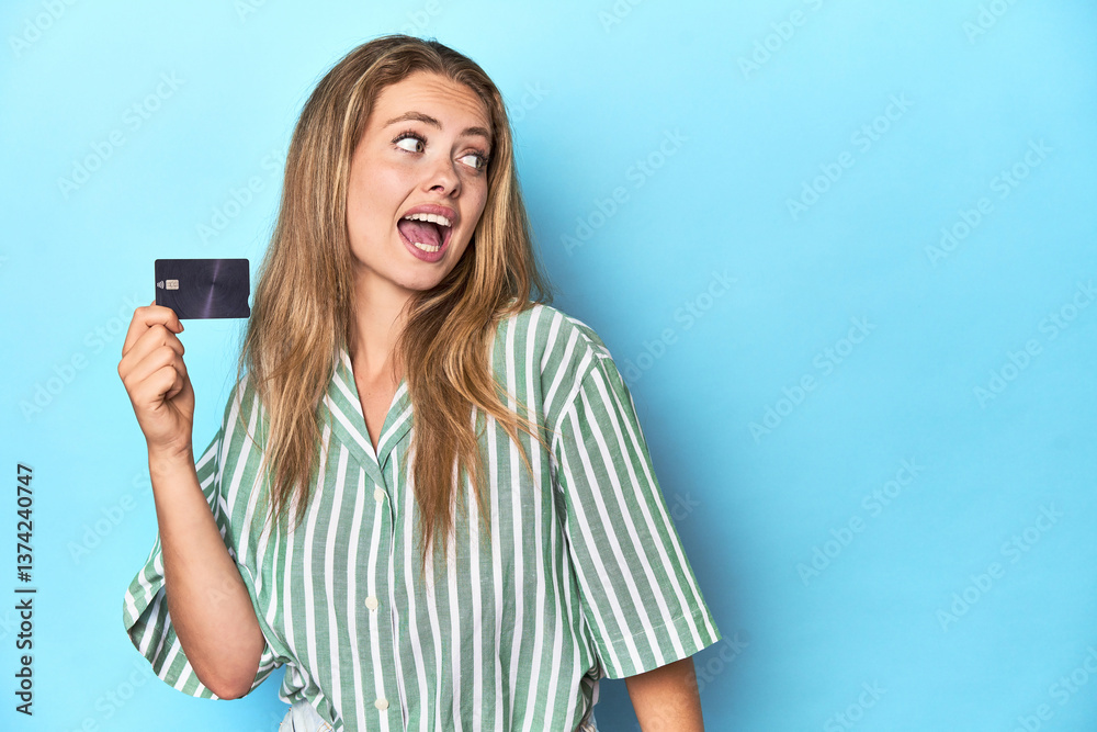 Young blonde with a credit card in blue studio points with thumb finger away, laughing and carefree. Young blonde with a credit card in blue studio points with thumb finger away, laughing and carefree.