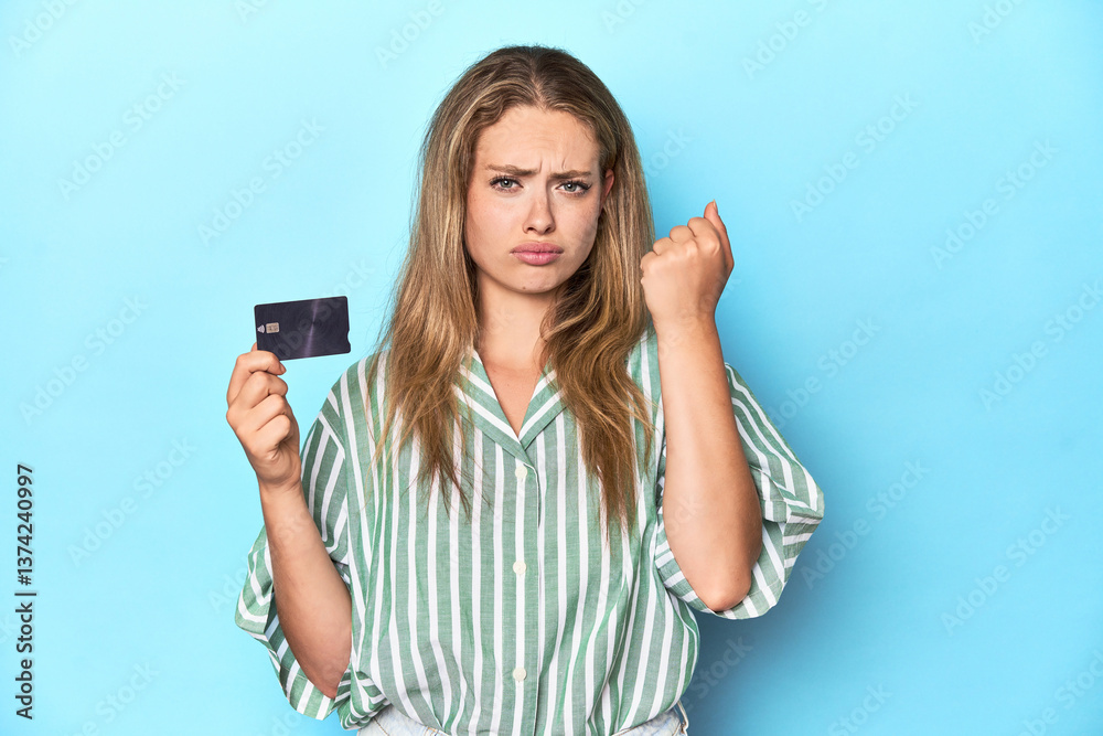 Young blonde with a credit card in blue studio showing fist to camera, aggressive facial expression.