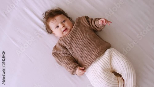 Smiling baby girl wearing warm knitted sweater and pant lying in bed over white blanket. Mother helps put on clothes.  