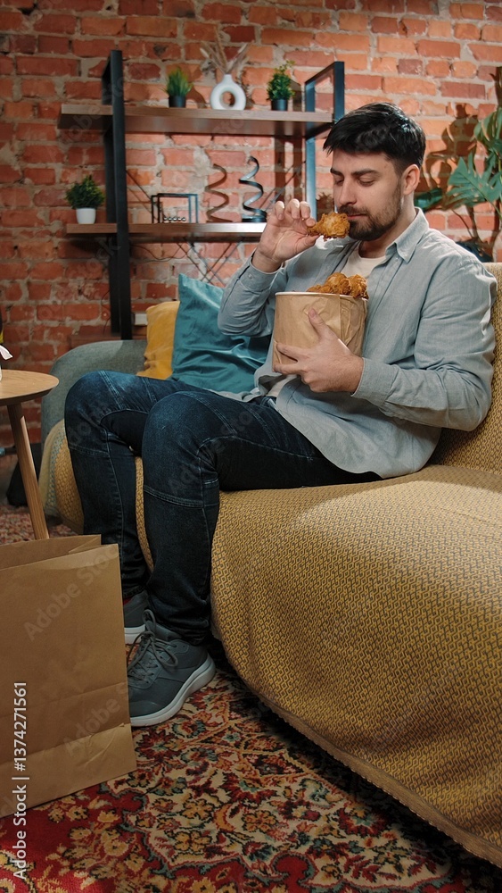 Fototapeta premium Man spending time at home with fast food delivery, sits on couch watching tv, dipping fried chicken leg in sauce and biting it, enjoying the taste.