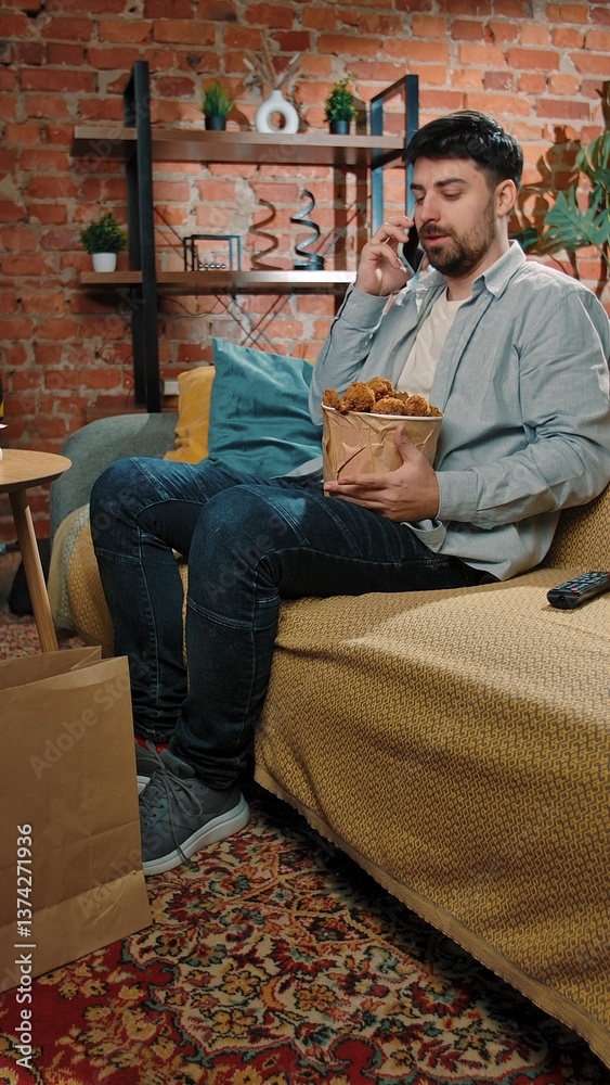 Fototapeta premium Man spending time at home with fast food delivery, sits on couch eating friend chicken leg and talking on smartphone.