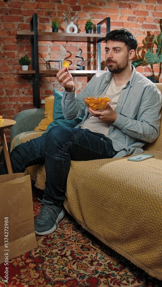 Fototapeta premium Bearded man spending time at home with fast food delivery, sits on couch watching tv and eating potato chips from a bowl.