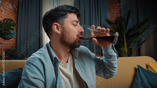 Φωτογραφία Bearded man spending time at home with fast food delivery, sits on couch and drinking cola from a large glass, taking a sip and enjoying the taste